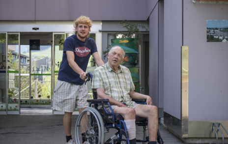 Ein junger Mann schiebt einen älteren Mann im Rollstuhl. Beide Männer sehen fröhlich aus.