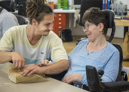 eine Klientin im Rollstuhl schneidet im Atelier von Viv Imboden ein Stück Jute mit einer Schere, unterstützt von einem Betreuer