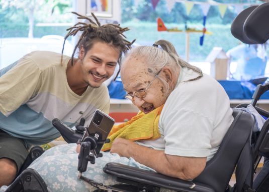 Ein Klientin im Rollstuhl und ein Betreuer schauen gemeinsam lachend auf ein Handy